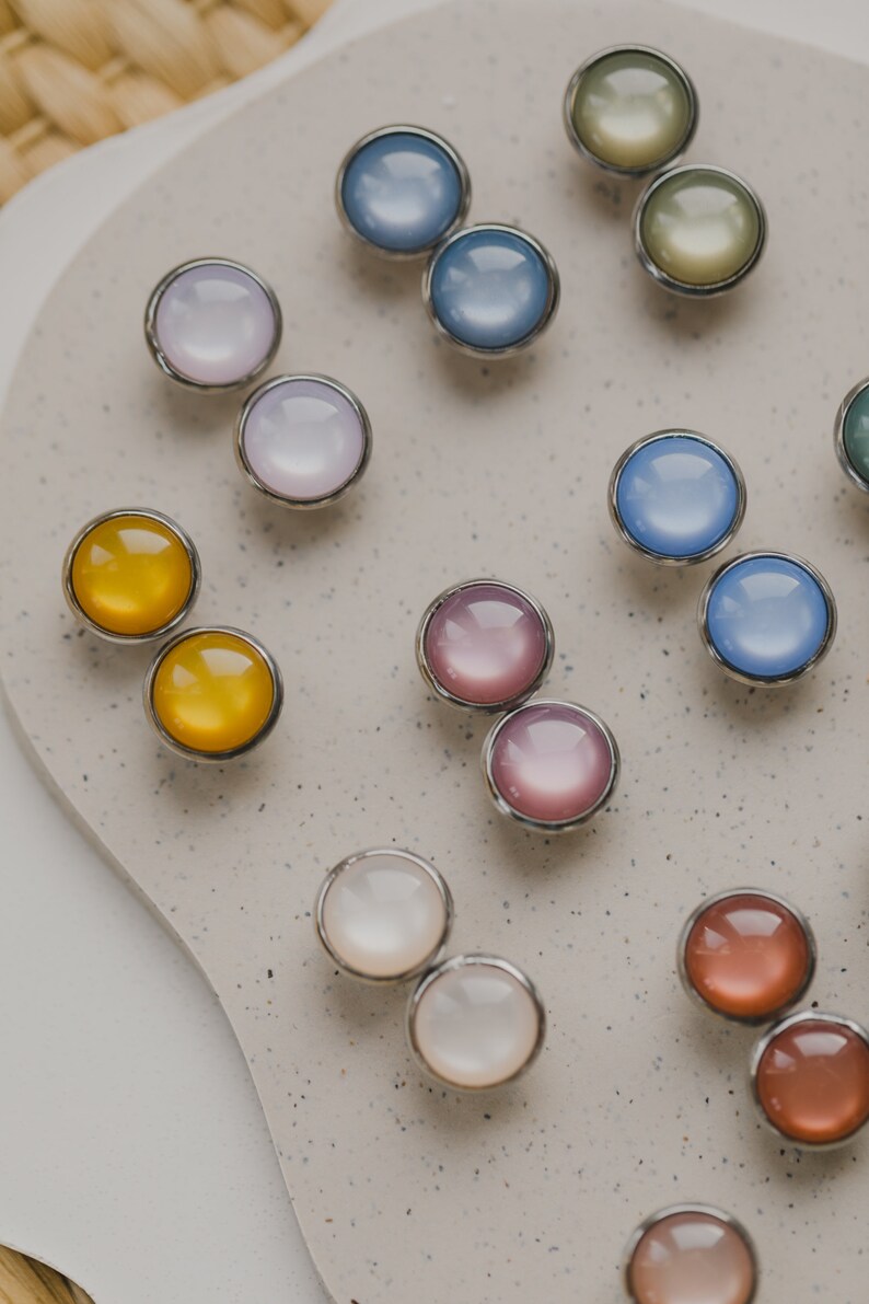 Op de afbeelding: Assortiment van ronde glazen cabochons in verschillende kleuren met zilveren instellingen. De cabochons zijn in paren gerangschikt op een lichtgrijs oppervlak.
