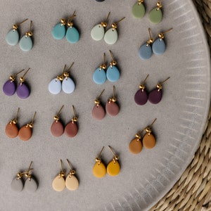 May include: A collection of 20 pairs of handmade ceramic earrings in various colors, including gray, blue, green, brown, purple, and yellow. The earrings are displayed on a gray plate with a textured surface.