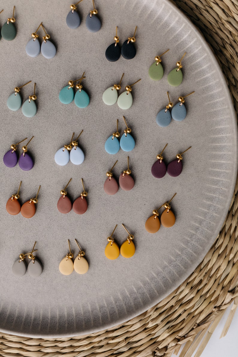 May include: A collection of 20 pairs of handmade ceramic earrings in various colors, including gray, blue, green, brown, purple, and yellow. The earrings are displayed on a gray plate with a textured surface.