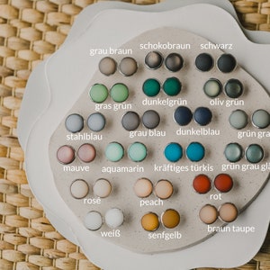 Op de afbeelding: Een display met 18 paar ronde, kleurrijke oorbellen op een witte achtergrond. De oorbellen zijn in een cirkel gerangschikt, waarbij elk paar is gelabeld met een Duits woord dat de kleur beschrijft.