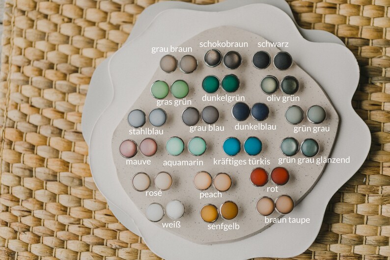 Op de afbeelding: Een display met 18 paar ronde, kleurrijke oorbellen op een witte achtergrond. De oorbellen zijn in een cirkel gerangschikt, waarbij elk paar is gelabeld met een Duits woord dat de kleur beschrijft.