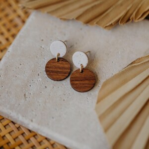 May include: A pair of earrings with white and brown round wooden discs. The earrings are on a light gray surface.
