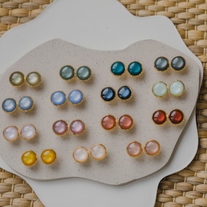 May include: A collection of 10 pairs of round, colorful, iridescent earrings displayed on a white, textured surface. The earrings are made of gold metal and have a variety of colors, including blue, green, pink, and yellow.