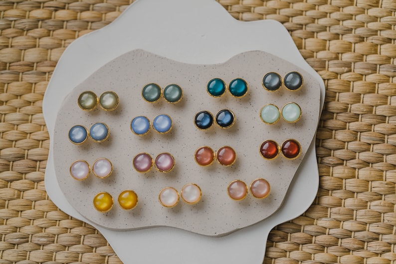 May include: A collection of 10 pairs of round, colorful, iridescent earrings displayed on a white, textured surface. The earrings are made of gold metal and have a variety of colors, including blue, green, pink, and yellow.