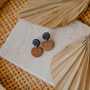 May include: A pair of earrings with black and brown round discs. The earrings are on a light brown surface with a dried palm leaf in the background.