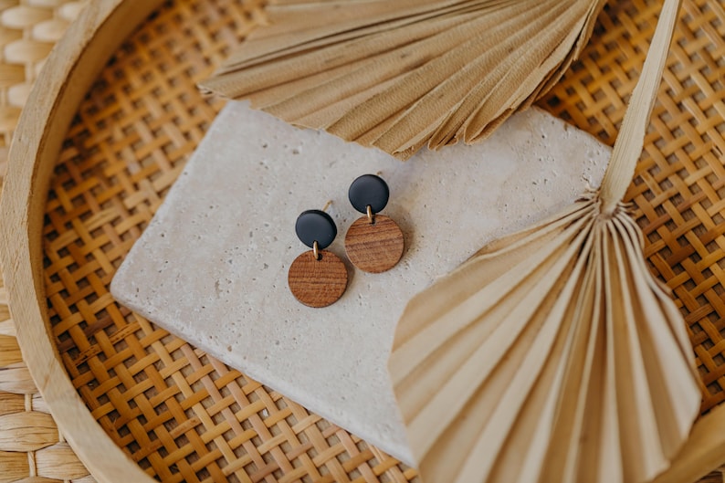 May include: A pair of earrings with black and brown round discs. The earrings are on a light brown surface with a dried palm leaf in the background.