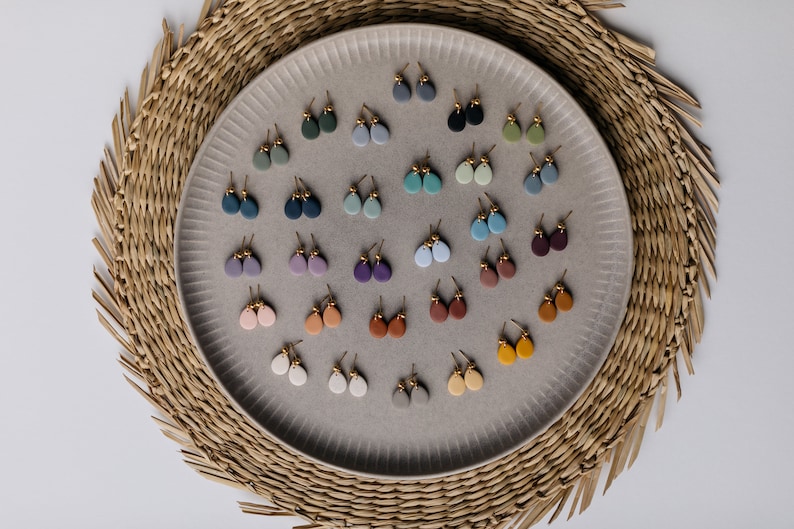 May include: A collection of 24 pairs of small, teardrop-shaped clay earrings in various colors, displayed on a gray plate. The earrings are arranged in rows and are all the same size and shape.