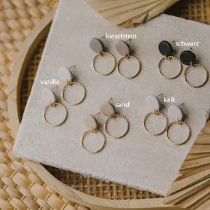 May include: Six pairs of gold hoop earrings with different colored clay discs. The colors are: vanille, kieselstein, schwarz, kalk, sand.