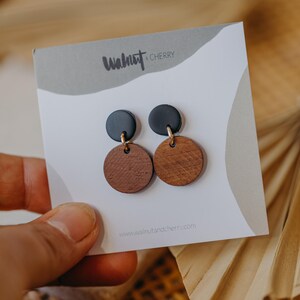 May include: A pair of earrings with black and brown round wooden discs. The earrings are on a white card with the text "Walnut & Cherry" and "www.walnutandcherry.com".