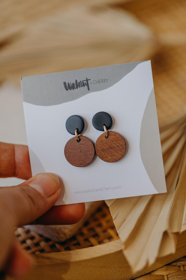 May include: A pair of earrings with black and brown round wooden discs. The earrings are on a white card with the text "Walnut & Cherry" and "www.walnutandcherry.com".