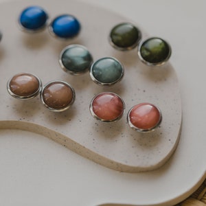 May include: A collection of colorful, round, gemstone earrings with silver settings. The earrings are displayed on a white surface.