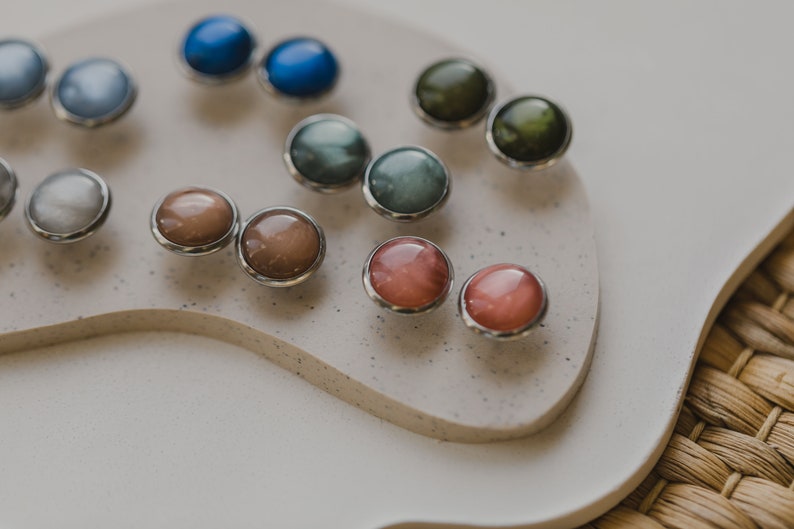 May include: A collection of colorful, round, gemstone earrings with silver settings. The earrings are displayed on a white surface.