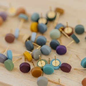 May include: A collection of colorful round stud earrings with gold posts. The earrings are scattered on a wooden surface.