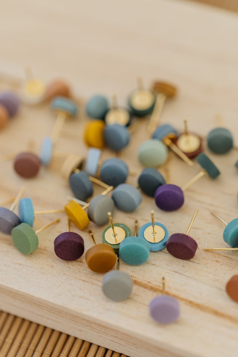 May include: A collection of colorful round stud earrings with gold posts. The earrings are scattered on a wooden surface.