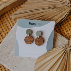 May include: A pair of wooden and green clay earrings with gold accents. The earrings are displayed on a white card with the text "Walnut & Cherry".