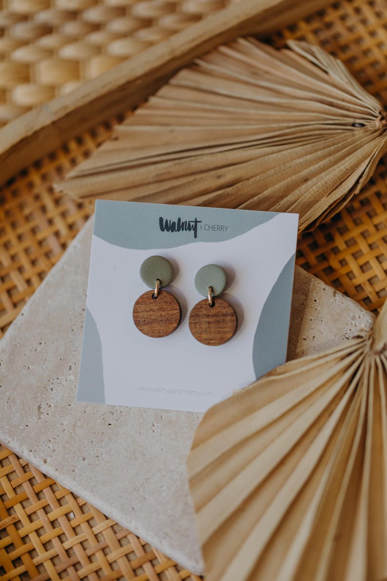 May include: A pair of wooden and green clay earrings with gold accents. The earrings are displayed on a white card with the text "Walnut & Cherry".