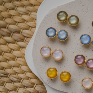 May include: Assortment of colorful round earrings with gold settings displayed on a white and beige surface. The earrings are arranged in pairs and feature various colors, including blue, pink, yellow, and green.