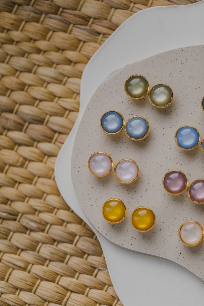 May include: Assortment of colorful round earrings with gold settings displayed on a white and beige surface. The earrings are arranged in pairs and feature various colors, including blue, pink, yellow, and green.