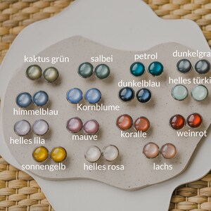 Op de afbeelding: Een display met 14 paar kleine ronde oorbellen in verschillende kleuren, waaronder groen, blauw, grijs, roze, rood, geel en wit. Elk paar is gelabeld met een Duits woord dat de kleur beschrijft.
