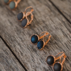 May include: Four pairs of hoop earrings with different colored stones. The earrings are on a wooden surface. The stones are black, green, and blue.