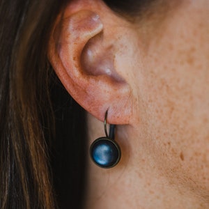 May include: A pair of dark blue button earrings with a bronze hook. The earrings are hanging from the earlobe of a person.