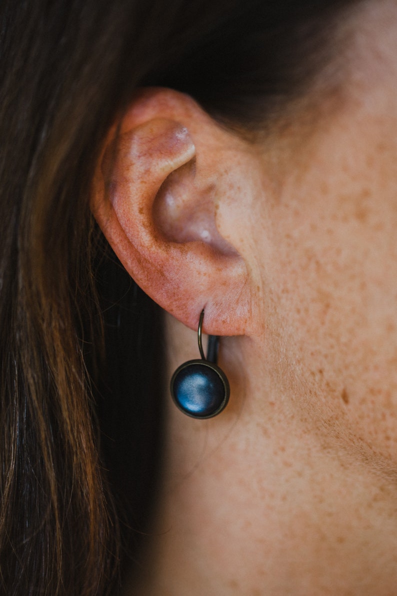May include: A pair of dark blue button earrings with a bronze hook. The earrings are hanging from the earlobe of a person.