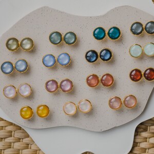 May include: A collection of 12 pairs of round, colorful earrings displayed on a white surface. The earrings are made of gold metal and have a variety of colors, including blue, green, pink, and white.