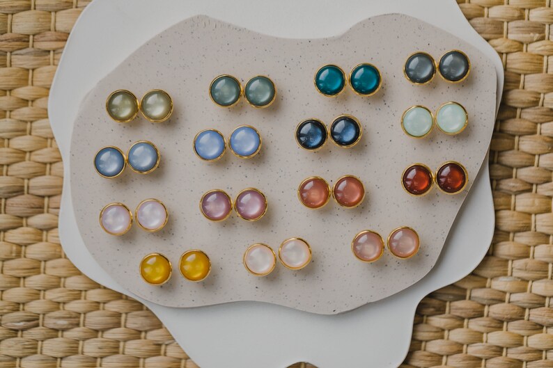 May include: A collection of 12 pairs of round, colorful earrings displayed on a white surface. The earrings are made of gold metal and have a variety of colors, including blue, green, pink, and white.