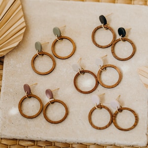 May include: Six pairs of wooden hoop earrings with different colored accents. The earrings are displayed on a light gray surface.