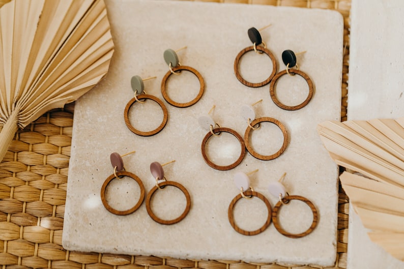 May include: Six pairs of wooden hoop earrings with different colored accents. The earrings are displayed on a light gray surface.