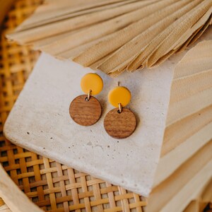 Puede incluir: Un par de pendientes con un c&iacute;rculo de madera y un c&iacute;rculo amarillo en la parte superior. Los pendientes est&aacute;n sobre una superficie blanca con un fondo tejido.