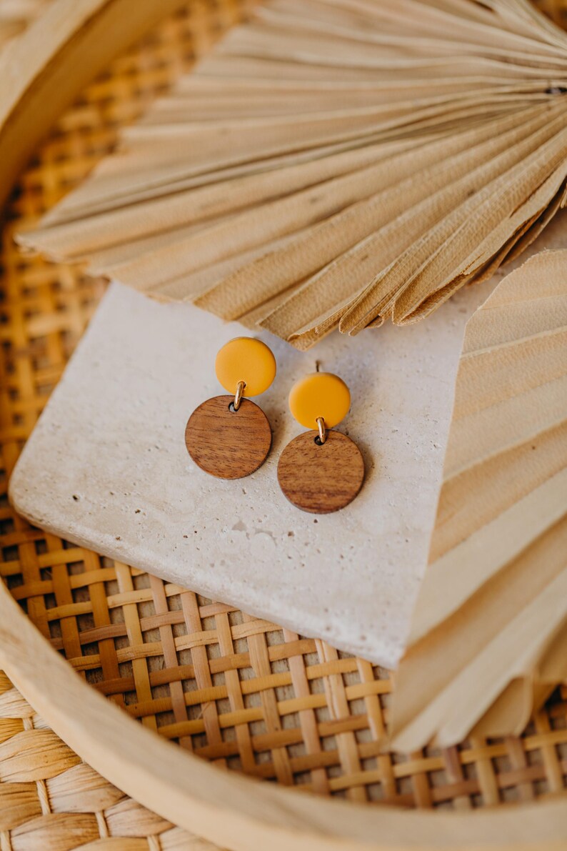 Puede incluir: Un par de pendientes con un c&iacute;rculo de madera y un c&iacute;rculo amarillo en la parte superior. Los pendientes est&aacute;n sobre una superficie blanca con un fondo tejido.