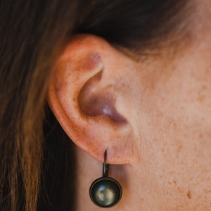 May include: A close-up of a person's ear with a small, round, dark grey earring. The earring has a simple, minimalist design.