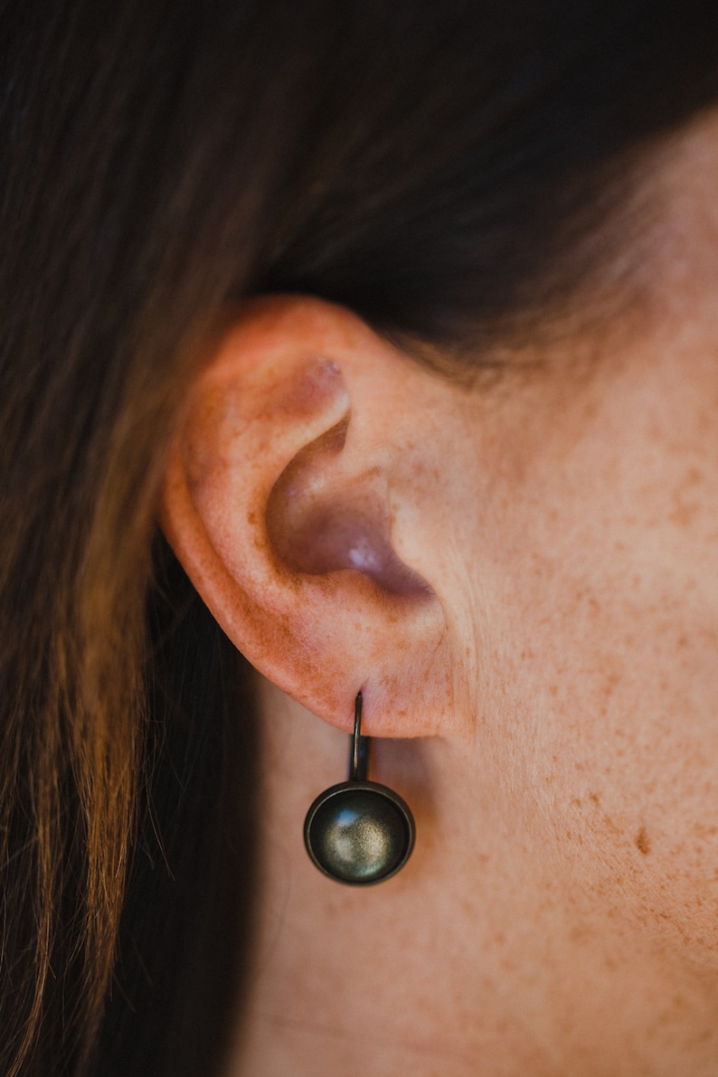 May include: A close-up of a person's ear with a small, round, dark grey earring. The earring has a simple, minimalist design.