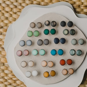Op de afbeelding: Een display met 24 paar kleine, ronde, kleurrijke oorbellen. De oorbellen zijn gerangschikt in rijen op een wit, getextureerd oppervlak. De oorbellen zijn gemaakt van verschillende materialen, waaronder glas, metaal en plastic.