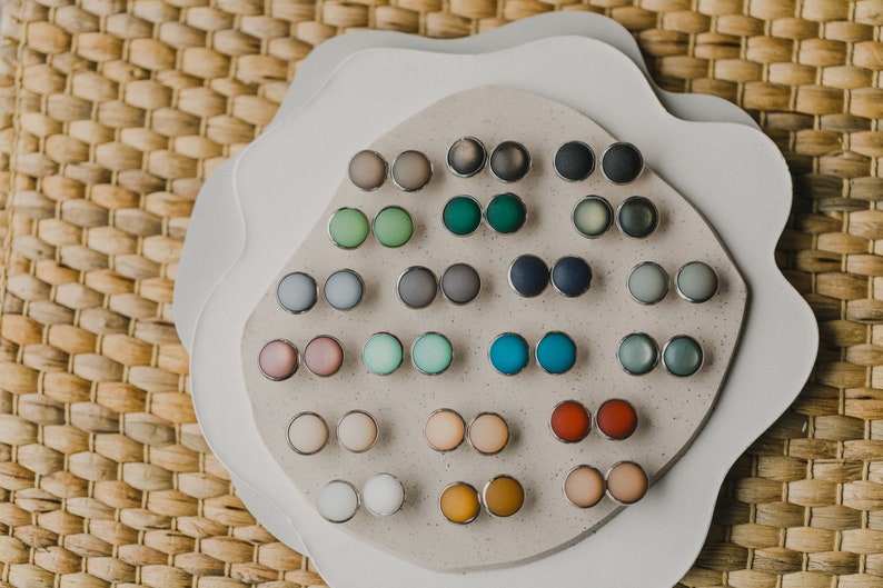 Op de afbeelding: Een display met 24 paar kleine, ronde, kleurrijke oorbellen. De oorbellen zijn gerangschikt in rijen op een wit, getextureerd oppervlak. De oorbellen zijn gemaakt van verschillende materialen, waaronder glas, metaal en plastic.