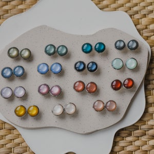 Op de afbeelding: Een verzameling van 10 paar ronde, iriserende oorbellen die worden getoond op een wit, getextureerd oppervlak. De oorbellen zijn in verschillende kleuren, waaronder blauw, groen, paars, roze en geel.