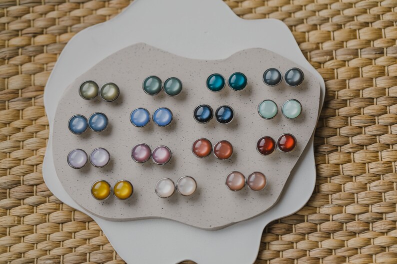Op de afbeelding: Een verzameling van 10 paar ronde, iriserende oorbellen die worden getoond op een wit, getextureerd oppervlak. De oorbellen zijn in verschillende kleuren, waaronder blauw, groen, paars, roze en geel.