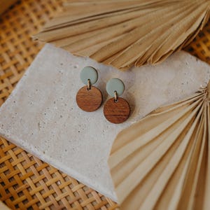 May include: A pair of wooden and green clay earrings. The earrings are round and have a simple design. They are displayed on a white surface with a woven background.