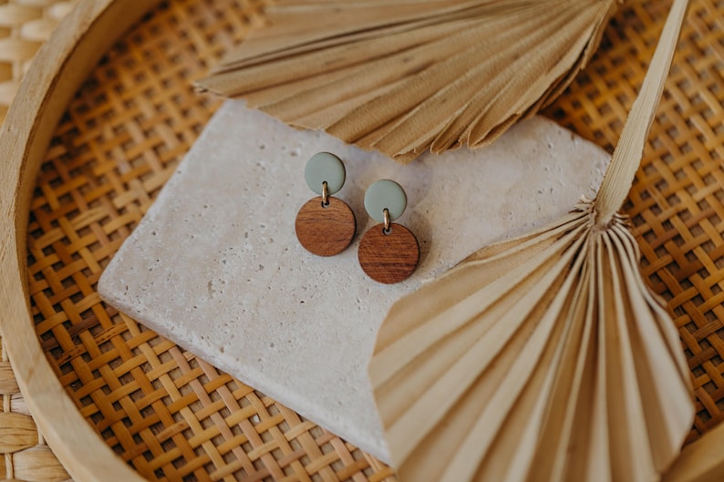 May include: A pair of wooden and green clay earrings. The earrings are round and have a simple design. They are displayed on a white surface with a woven background.