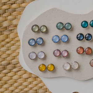 Op de afbeelding: Een verzameling van 10 paar kleine, ronde, kleurrijke oorbellen die op een wit en beige oppervlak worden getoond. De oorbellen zijn gemaakt van metaal en hebben een verscheidenheid aan kleuren, waaronder blauw, groen, roze, geel en wit.