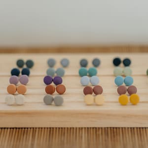 May include: A set of 18 pairs of small, round, colorful earrings displayed on a wooden surface. The earrings are arranged in rows of two, with each pair featuring a different color.