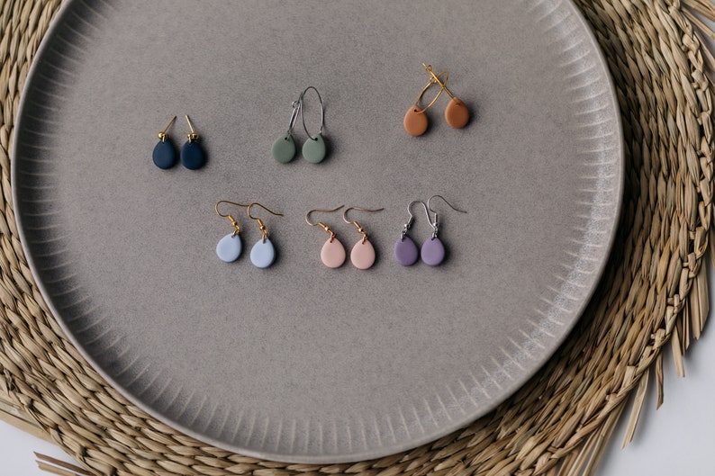 May include: A set of 10 dangle earrings with different colored clay teardrop shapes. The earrings are displayed on a gray plate with a woven texture.