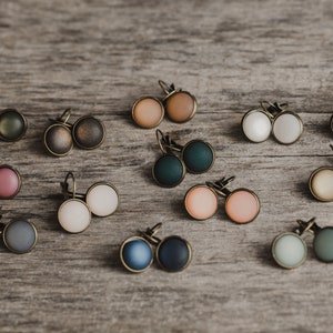 May include: Assortment of 10 pairs of earrings with various colours and finishes. The earrings are displayed on a wooden surface. The colours include green, brown, pink, white, blue, and beige.