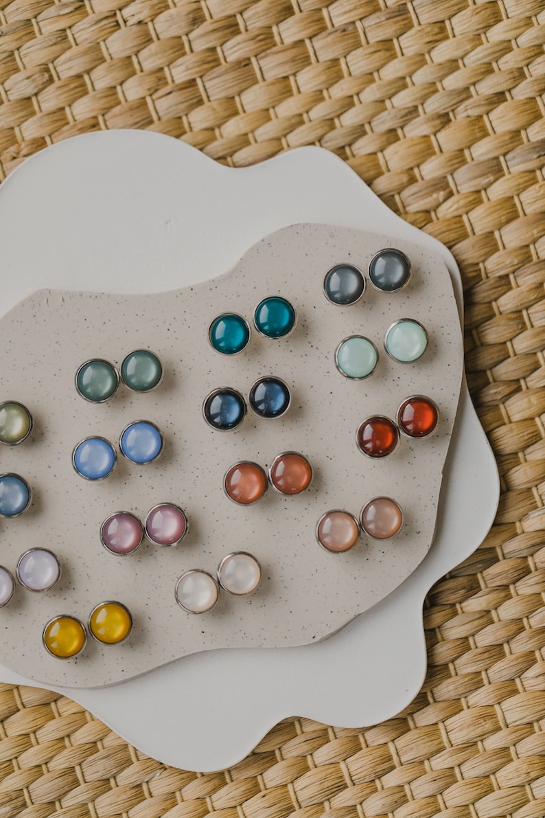 Op de afbeelding: Een verzameling van 10 paar regenboogkleurige ronde oorbelletjes, gepresenteerd op een wit en beige oppervlak. De oorbelletjes zijn in rijen gerangschikt en tonen een verscheidenheid aan kleuren, waaronder blauw, groen, roze, rood en geel.