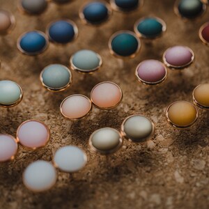 Pu&ograve; includere: Assortimento di orecchini rotondi colorati con montature dorate. Gli orecchini sono esposti su una superficie di sughero.