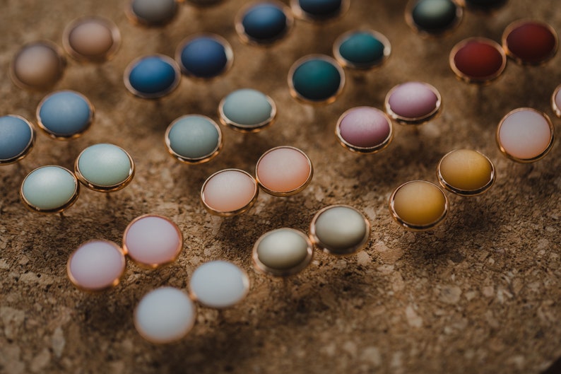 Pu&ograve; includere: Assortimento di orecchini rotondi colorati con montature dorate. Gli orecchini sono esposti su una superficie di sughero.
