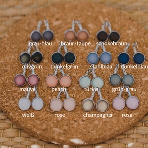 May include: A set of 16 pairs of earrings with round glass cabochons in various colors, including gray, blue, green, brown, peach, pink, white, and turquoise. The earrings are displayed on a cork board with the color names written below each pair.