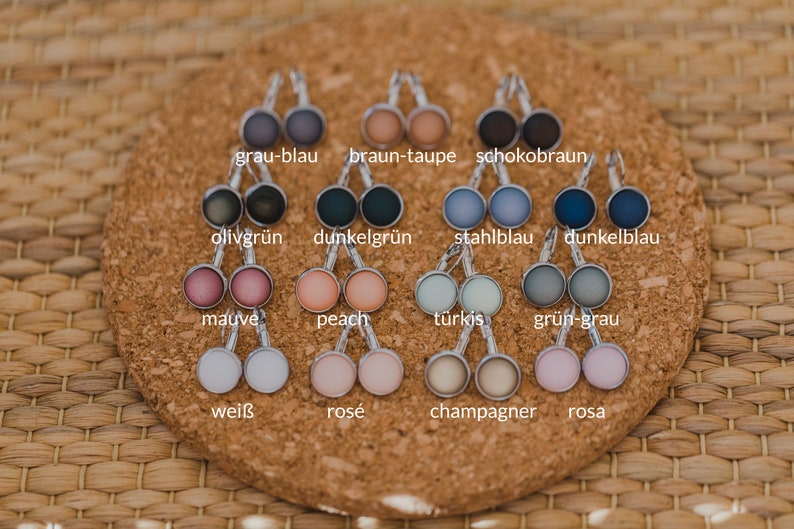 May include: A set of 16 pairs of earrings with round glass cabochons in various colors, including gray, blue, green, brown, peach, pink, white, and turquoise. The earrings are displayed on a cork board with the color names written below each pair.