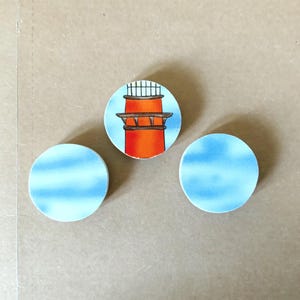 May include: Three round ceramic tiles. One tile features a lighthouse design with an orange tower and a blue sky background. The other two tiles have a blue sky design.